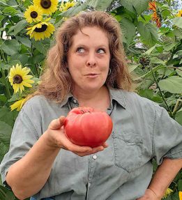 a farmer in front of sunflowers holds a large red heirloom tomato in one hand while looking playfully to one side