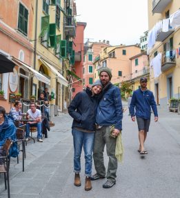 Two smiling people stand on a busy village street in Italy; they are flanked by pedestrians and people eating at an outdoor cafe and colored buildings rise behind them