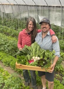 Two farmers hold a wooden box laden with fresh produce while standing in a hoophouse full of plants