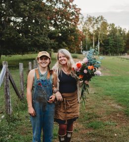 Two farmers at Winterberry Farm stand on a green field. One holds an autumnal bouquet of eucalyptus and dahlias.