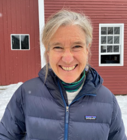 MOFGA board member, Martha Leggat, stands in front of a red barn.